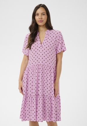 Kobieta o długich ciemnych włosach, ubrana w różową sukienkę na krótkie rękawy w brązowe kropki, stojąca na białym tle.