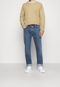 Hombre con suéter beige, jeans azules remangados en los puños, zapatillas blancas, cinturón marrón, de pie contra un fondo blanco liso.