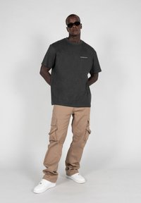 Graues T-Shirt mit einem Logo, beiges Cargo-Hose mit mehreren Taschen und weiße Sneaker. Der Model trägt eine Sonnenbrille und steht mit den Händen hinter dem Rücken.