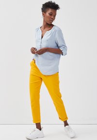 Chemise à manches longues bleu clair avec un col en V et une poche poitrine, associée à un pantalon à revers jaune vif et des baskets blanches.