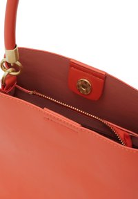 Korallinvärinen nahkakäsilaukku, jossa on sileä pinta, kullanvärinen nepparilukitus ja sisällä vetoketjutasku. Kahva on myös korallinvärinen.