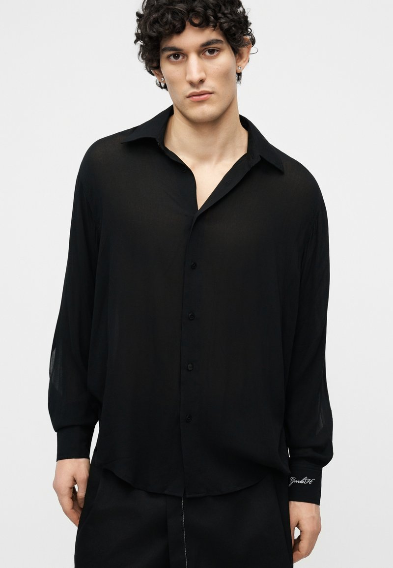 Young man with curly hair wearing a loose black button-up shirt and black pants, standing against a plain light gray background.