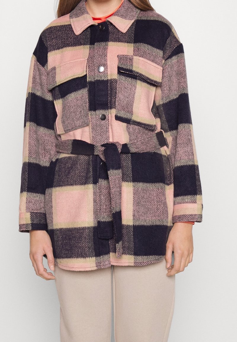 Femme portant une veste à carreaux oversize avec ceinture et grandes poches en rose, beige et noir, associée à un pantalon beige clair.