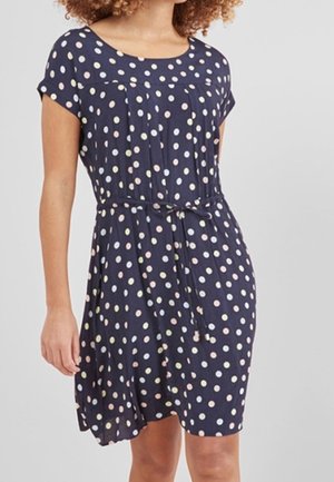 Kobieta ubrana w granatową sukienkę do kolan z krótkimi rękawami, okrągłym dekoltem, pastelowymi groszkami oraz przewiązanym paskiem w pasie.