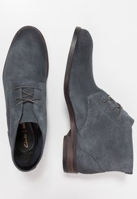 Bottines en daim gris avec un devant à lacets, bouts ronds et talons en bois. Présente des coutures visibles et une texture lisse.
