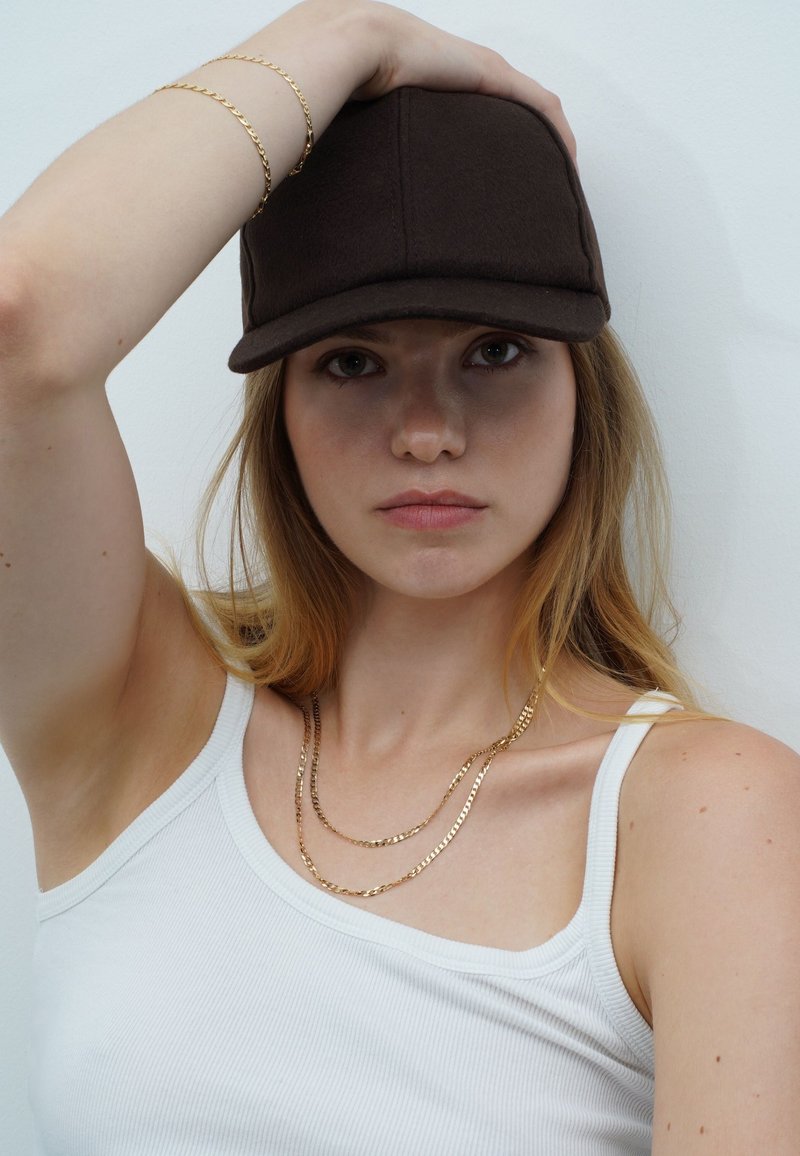 Braune Stoffbasecap, geschichtete goldene Kettenhalsketten und ein weißes geripptes Tanktop. Das Haar des Models ist glatt und wellig.