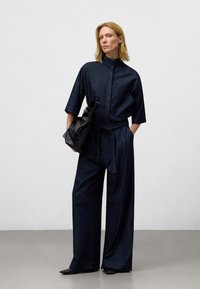 Tuta in denim scuro con colletto alto, maniche corte e vita cintrata. Abbinata a una borsa nera e scarpe con punta.