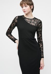 Noor naine mustas, keha lähedases kleidis, millel on läbipaistvad pikad varrukad ja lilleline sädelus, poseerib heleda ühtlase tausta ees.