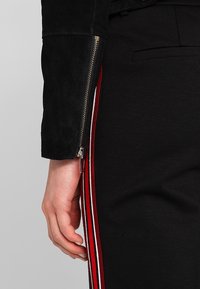 Ruka odpočívající vedle černých kalhot s červenými a bílými bočními pruhy, na sobě má černou bundu se stříbrným zipem na rukávu.