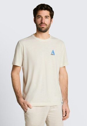 Mann mit dunklem Haar und Bart, der ein beiges T-Shirt mit kleinem blauem Segelboot-Logo und hell gestreifte Hosen trägt, steht mit einer Hand in der Tasche.