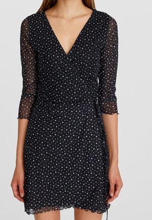 Frau trägt ein schwarzes Wickelkleid mit weißen Polka-Dots, transparenten Dreiviertelärmeln und einem gewellten Saum.
