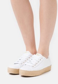 Sneakers bianche in tela con suola in corda di juta, caratterizzate da occhielli dorati, lacci e design con punta rotonda. Tessitura liscia e stile minimalista.