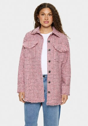 Roze en zwarte geblokte jas met een kraag, twee borstzakken, knoopsluiting en een rafelzoom. Draagt over een witte top en blauwe spijkerbroek.