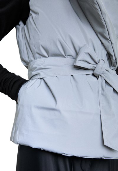 Grå vadderad väst med bälte i midjan, som har en slät textur och mjukt material. Västen har en åtsittande design och dekorativ knyt-detail.