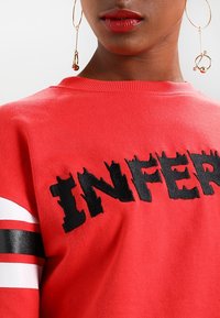 Sudadera roja con texto negro envejecido que dice "INFER". Presenta acentos de rayas blancas y negras en las mangas. Textura de tela suave.