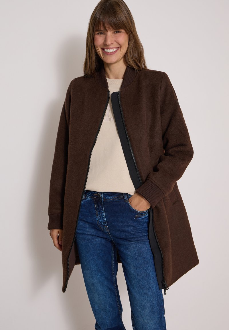 Femme souriante portant un manteau marron zippé sur un haut beige clair et un jean bleu, une main dans la poche, sur un fond uni.