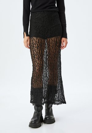 Maxifalda de encaje negro con un diseño transparente, que presenta un patrón floral, pantalones cortos forrados debajo y un dobladillo recto. Combinada con botas negras.