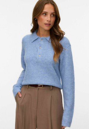 Femme aux longs cheveux bruns portant un pull duveteux bleu clair avec des boutons et un pantalon beige taille haute, les mains dans les poches.