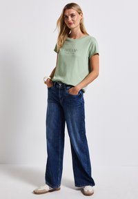 Světl zelené tričko s krátkým rukávem a nápisem "NĚKONEČNÝ SEN", spárované s tmavě modrými širokými džínami a bílými teniskami. Jednoduchý design, uvolněný střih.