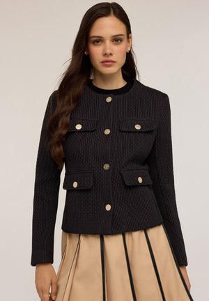 Chaqueta negra texturizada con un diseño ajustado, que presenta dos bolsillos en el pecho y botones dorados. Combinada con una falda beige plisada.