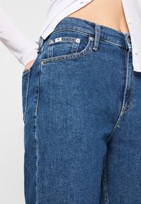 Tmavě modré džíny z denimu se rovnými nohavicemi, se standardním zapínáním na knoflíky, pěti kapsami a viditelnými švy.