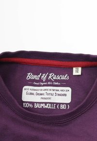 Lila bomullströja med en röd inre krage, med en etikett som visar varumärket, skötselråd och detaljer om ekologisk certifiering.