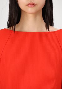 Gros plan d'une personne portant un haut côtelé rouge vif avec un large décolleté carré montrant le cou et les lèvres.