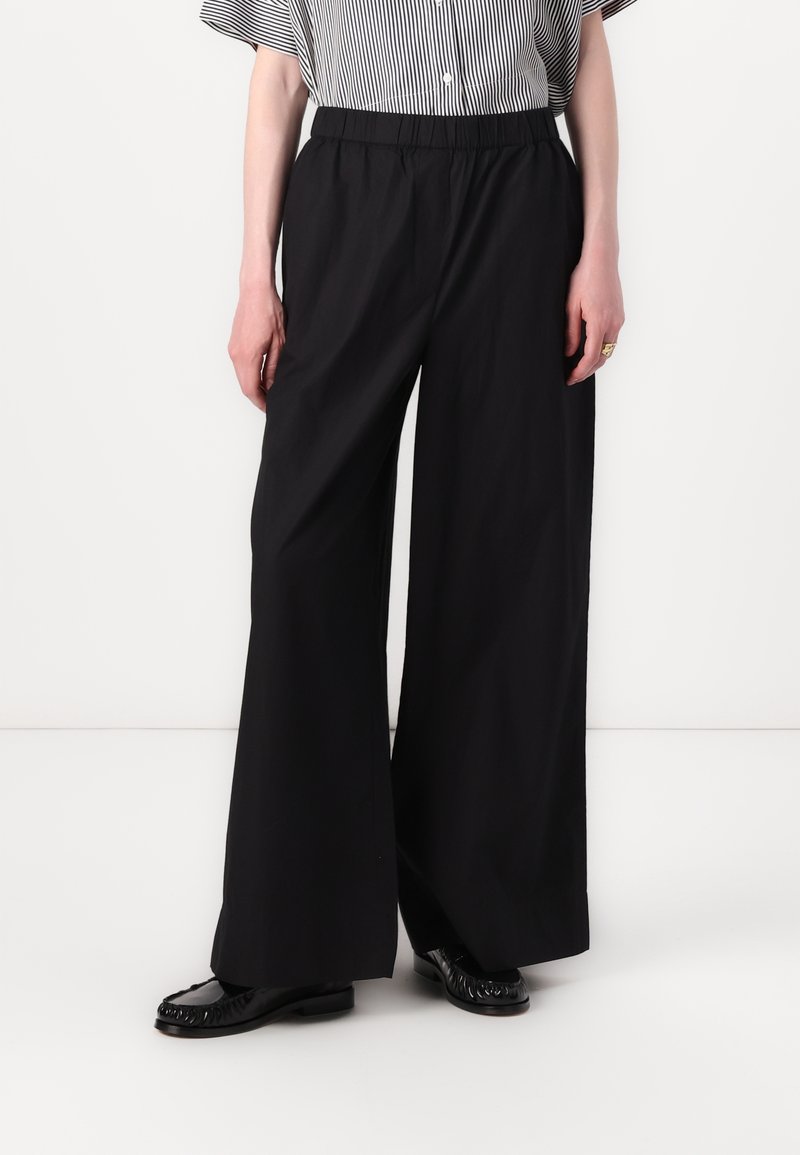 Personne portant un pantalon noir à jambes larges, des mocassins noirs et un haut à manches courtes rayé noir et blanc rentré, debout devant un fond blanc.