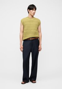 Un homme porte un haut en maille vert sans manches, un pantalon large à rayures sombres, une ceinture marron et des sandales foncées, sur fond blanc.