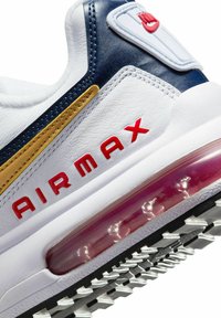 Sneaker blanc avec dessus en cuir, accents bleu marine, bande dorée et texte "AIR MAX" rouge. Présente un amorti à air transparent et une semelle noire texturée.