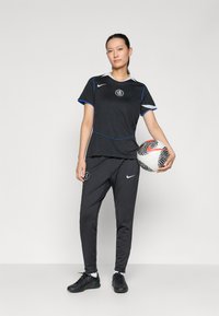 Svart fotballantrekk med blå aksenter. T-skjorte med korte ermer og et logo. Løse bukser. Holder en svart og hvit mønstrete fotball.