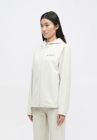 Veste zippée beige clair avec capuche, arborant le logo "Terrex" sur la poitrine. Tissu lisse et coupe décontractée. Design simple avec des accents minimalistes.