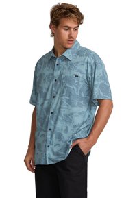 Young man wearing a loose, short-sleeve, button-up blue patterned shirt with one hand in the pocket of black pants, looking down to side.