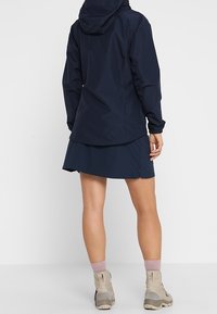 Mörkblå vattentät jacka med huva, elastiska ärmar, i kombination med en matchande kjol. Bärs med beige ankelskor och rosa strumpor. Slät tygtextur.