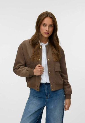 Jeune femme aux longs cheveux bruns porte une veste marron boutonnée par-dessus une chemise blanche et un jean bleu, debout devant un fond uni.