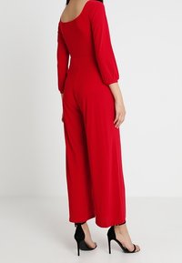 Mono rojo con escote en la espalda, mangas largas y piernas anchas de corte holgado. Hecho de una tela suave, presenta una cintura ceñida, combinado con tacones negros.