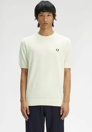 Uomo che indossa un maglione bianco semplice a maniche corte con un piccolo logo di alloro nero sul petto e pantaloni scuri, in piedi davanti a uno sfondo bianco.