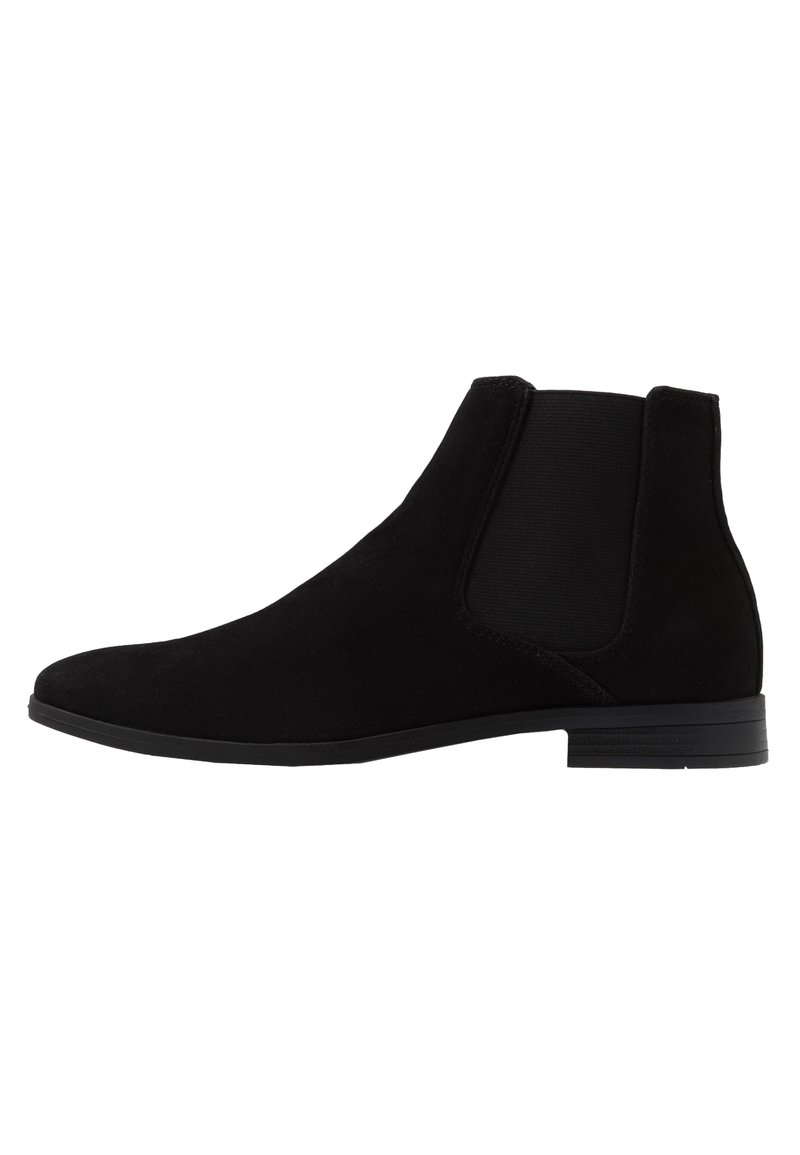 Botas de tornozelo em camurça preta com painéis laterais elásticos, bico arredondado e sola de borracha baixa e flat, exibindo um design minimalista.
