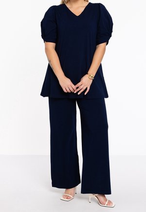 Kvinde iført marineblå bukser med vide ben og matchende kort top med puffede ærmer, beige højhælede sandaler og et guldarmbånd på venstre håndled.