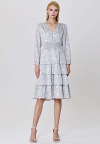 Jeune femme portant une robe à manches longues à motif floral, avec une taille froncée et des chaussures à talons avec strap blanc, se tenant devant un fond uni.