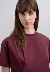 Bordeaux kleurige top met korte mouwen, gemaakt van zachte katoen. Heeft een losse pasvorm en een subtiel plooidetail aan de voorkant. Soepele textuur, ronde hals.