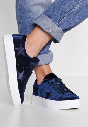 Stopy w ciemnoniebieskich welurowych trampkach z wzorami gwiazd i grubymi białymi podeszwami, w połączeniu z podwiniętymi niebieskimi dżinsami.