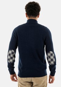 Marineblauwe trui van zacht materiaal, met geruite elleboogpatches. Hoge kraag en ribgebreide manchetten. Getailleerde pasvorm met een gladde textuur.