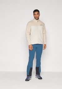 Fleecejakke i beige med lynlås og høj krave, parret med blå bukser med sorte detaljer og åndbart stof.