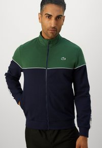 Jaqueta masculina com fecho zip em azul escuro e verde. Apresenta um colarinho alto, blocos de cor contrastantes e um logotipo discreto no peito. Feita de um tecido suave.