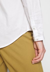 Camicia bianca con bottoni, dal tessuto morbido e orlo curvo, abbinata a pantaloni color giallo senape. La camicia presenta polsini con bottone.
