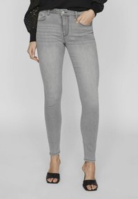 Jeans ajustados grises en un material de mezclilla elástica con una cintura de media altura, diseño de cinco bolsillos y acentos de costura, combinados con sandalias de tacón negras.