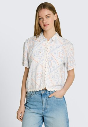 Frau trägt eine kurzärmelige Bluse mit Kragen, wellenförmigen Kanten und pastellfarbenem geometrischem Muster, kombiniert mit hoch taillierten hellblauen Jeans.
