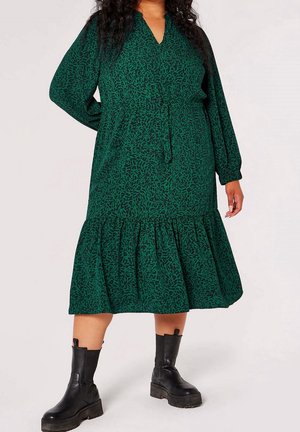 Frau trägt ein grünes, gemustertes Midi-Kleid mit langen Ärmeln, einem gerafften Saum und schwarzen Plateau-Stiefeletten, steht mit einer Hand hinter dem Rücken.