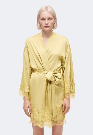 Femme blonde portant une robe en satin jaune pâle avec bordure en dentelle aux manches et à l'ourlet, nouée à la taille avec un nœud, debout devant un fond neutre.